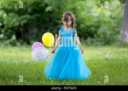 Hübsche kleine Blonde langhaarige Mädchen in schönen langen blauen Abendkleid hält bunte Luftballons in blühenden Feld auf verschwommen grünen Bäumen, backgro Stockfoto