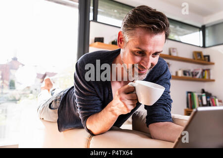 Portrait von lächelnden Mann mit Tasse Kaffee liegen auf der Rückenlehne der Couch mit Tablet Stockfoto