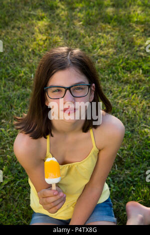 Portrait von Mädchen mit Brille sitzen auf einer Wiese Eis essen Lolly Stockfoto