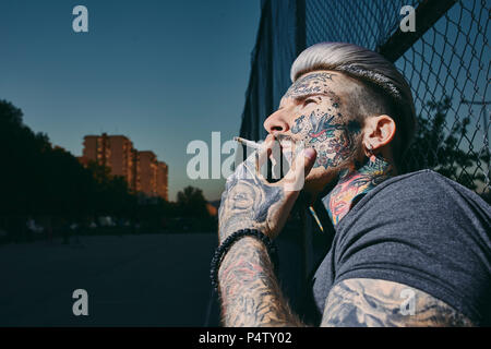 Portrait von tätowierten jungen Mann Rauchen einer Zigarette am Maschendrahtzaun Stockfoto