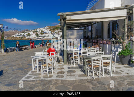 Mykonos, Griechenland Masse an der Taverne stil Fast Food Restaurant. Die Menschen essen auf Sitzgelegenheiten im Freien an Tischen mit weißen Stadt am Wasser im Zentrum von Manto Square. Stockfoto