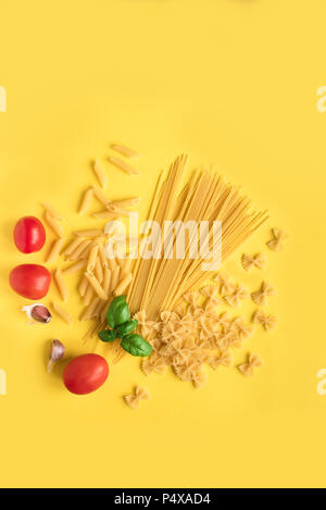 Verschiedene roh Rohe Nudeln mit Basilikum, Tomaten und Knoblauch auf gelben Hintergrund. Italienische Pasta: Penne, Farfalle, Spaghetti. Stockfoto