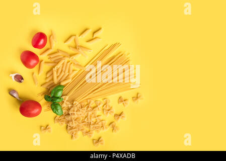 Verschiedene roh Rohe Nudeln mit Basilikum, Tomaten und Knoblauch auf gelben Hintergrund. Italienische Pasta: Penne, Farfalle, Spaghetti. Stockfoto