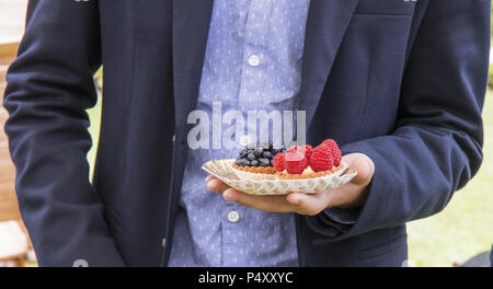 Ein Mann im Anzug hält eine Schüssel mit süßen Früchten Desserts. Gebäck - süße Tassen mit Himbeeren und das andere mit Heidelbeeren. Essen shopping dur Stockfoto