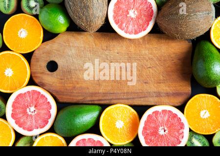 Flach. Ansicht von oben. Holzbrett mit in Scheiben geschnittenen bunten frische Früchte: Kiwi, Orange, Grapefruit und Mandarine. Kokosnüsse, Kalk und Avocado. Schwarz backgrou Stockfoto