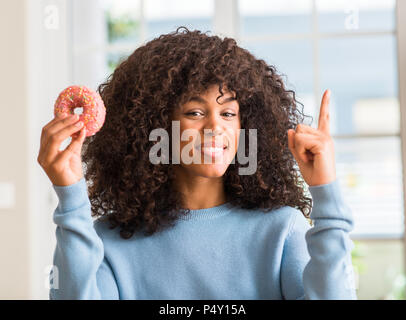 African American woman holding Donut zu Hause mit einer Idee oder Frage Zeigefinger mit glücklichen Gesicht überrascht, Nummer eins Stockfoto