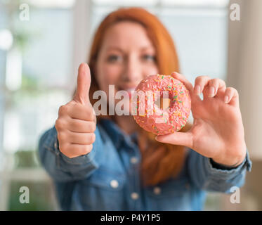 Rothaarige Frau mit Donut zu Hause glücklich mit dem großen Lächeln tun ok Zeichen, Daumen hoch, mit den Fingern, ausgezeichnete Zeichen Stockfoto