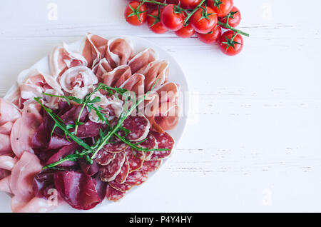 Kalt geräucherte Fleisch Platte mit Schweinefleisch, Schinken, Salami, Bresaola, Rucola und Tomaten cherry auf weißem Holz- Hintergrund. Wein Vorspeise gesetzt. Italienische fo Stockfoto