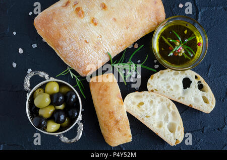Frisch gebackenes Brot mit traditionellen italienischen Brot ciabatta auf schwarzen Steintisch mit Rosmarin, Salz, Olivenöl und Oliven in Scheiben geschnitten. Italienisches Essen Konzept.