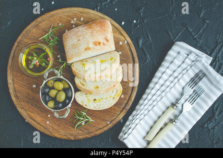 Mediterrane Snacks. Grüne und schwarze Oliven, Olivenöl, Kräuter und Scheiben ciabatta Brot auf Holzbrett über dunklen Stein Hintergrund. Italienisches Essen Stockfoto