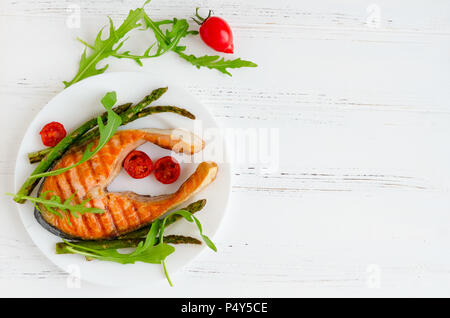 Gegrilltes Lachssteak mit Spargel und Tomaten cherry auf einem Teller mit Rucola auf weißem Holz- Hintergrund mit Platz für Text. Gesundes Mittagessen Konzept. Stockfoto