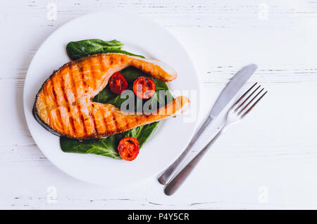 Gegrilltes Lachssteak mit Cherry Tomaten und Spinat auf einem Teller mit Messer und Gabel serviert auf weißem Holz- Hintergrund. Gesundes Mittagessen Konzept. Delici Stockfoto