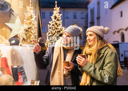 Junges Paar mit Tassen Glühwein auf Schaufenster in der Weihnachtszeit auf der Suche Stockfoto