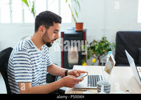 Junger Mann mit Laptop und Handy am Schreibtisch im Büro Stockfoto