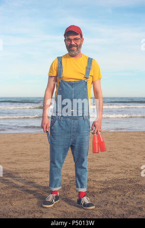 Portrait von lächelnden Mann in Latzhosen stehen auf dem Strand holding Flaschen Erfrischungsgetränke Stockfoto