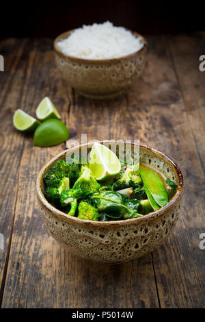 Green Thai curry mit Broccoli, Pak Choi, Zuckerschoten, Baby Spinat, Kalk und Schüssel Reis im Hintergrund Stockfoto