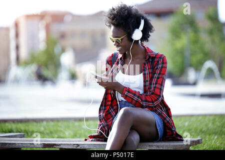 Lächelnde junge Frau sitzt auf der Bank im Stadtpark, Musik hören mit Kopfhörern Stockfoto