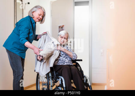 Frau kümmert sich um alte Frau im Rollstuhl setzen ihre Jacke auf Stockfoto