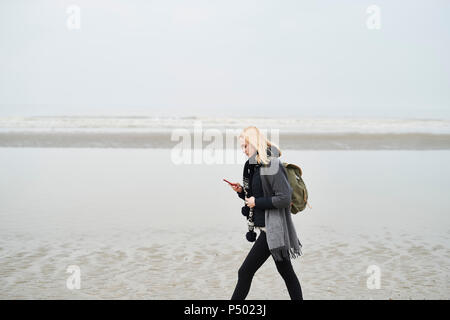 Niederlande, junge Frau mit Rucksack zu Fuß am Strand an der Zelle Telefon Stockfoto