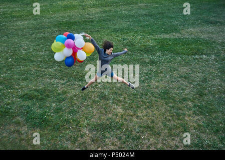 Glückliche junge Frau mit Bündel Luftballons springen in die Luft, auf einer Wiese Stockfoto
