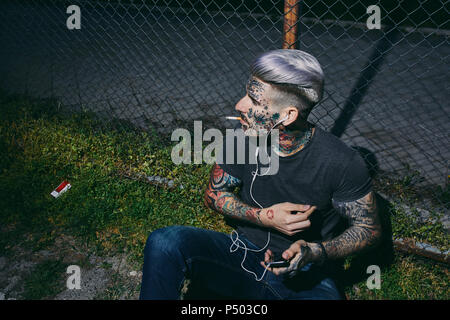 Tätowierten jungen Mann mit Ohrhörern und Smartphone das Rauchen einer Zigarette am Maschendrahtzaun Stockfoto