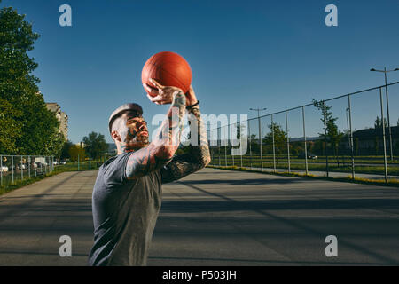 Tätowierte junge Mann werfen Basketball im Freien Gericht Stockfoto