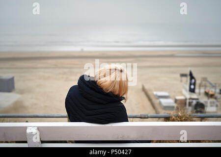 Rückansicht der blonde junge Frau sitzt auf der Bank mit Blick auf Strand und Meer im Winter Stockfoto