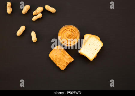 Toast, Erdnüsse ohne Schale und Schüssel von Erdnußbutter auf einem schwarzen Hintergrund, Ansicht von oben. Von oben. Stockfoto