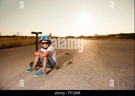 Nachdenklich, schöner kleiner Junge bei Sonnenbrillen und Helm sitzt auf einem Motorroller auf verlassenen Straße Stockfoto