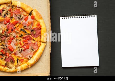 Frisch gebackene Pizza in einem Karton mit Notebook über schwarzen Hintergrund, Ansicht von oben. Von oben. Stockfoto