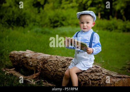 Ein litttle zwei Jahre alte Junge sitzt auf einem Baum und liest ein Buch. hildren Tag. Stockfoto