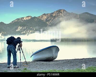 Fotograf mit Auge am Sucher ist unter Foto von See mit Alpen Berge im Hintergrund. Künstler arbeitet. Stockfoto