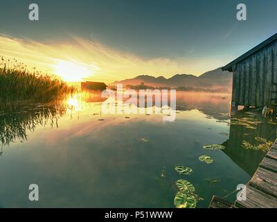 Romantischen sonnenaufgang Reflexion auf dem Kochelsee gegen Bootshäusern. Anfang Sommer morgen ansehen, Bayern, Deutschland Stockfoto