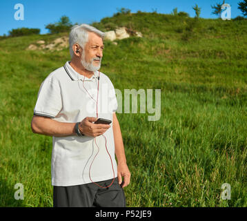 Alte Mann, Handys, die auf die Landschaft. Musik hören mit Kopfhörern. Tragen klassisches weißes Poloshirt mit dunkelblauem Streifen, schwarze Hose, Turnschuhe. Sport Lifestyle. Stockfoto