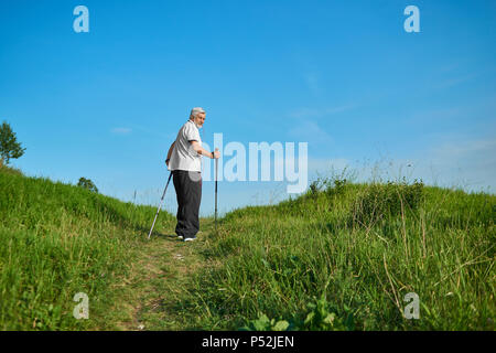 Man Walking im Feld mit Tracking Sticks. Gesättigt grünen Gras, strahlend blauen Himmel. Draußen Sport Aktivitäten. Tragen klassisches weißes Poloshirt mit dunkelblauem Streifen, Sportuhr. Gesunde Lebensweise. Stockfoto