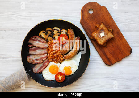 Ansicht von oben, englisches Frühstück in einer Pfanne mit Spiegelei, Speck, Bohnen, Würstchen und Toast auf einem weißen Holz- Hintergrund. Stockfoto