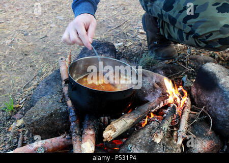 Kochen auf dem Scheiterhaufen. Rühren mit einem Löffel Suppe in Schwarz vom Ruß pot. Stockfoto