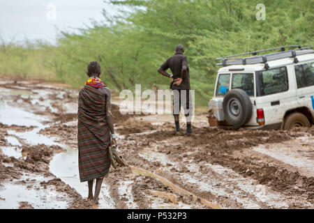 Kakuma, Kenia - ein Turkana Frau geht barfuß auf einer Schotterstraße nasse mit Regen. Im Hintergrund ein UN-Land Rover Festgefahren im Schlamm. Stockfoto