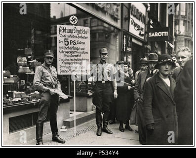 Das antijüdische rassistische Aufhetzungsbild der SA Sturmbleitung oder „Brownshirts“-Mitglieder der Sturmabteilung von Vintage 1930, die zum Boykott jüdischer Geschäfte in der Friedrichstraße, Berlin, aufriefen; 1. April 1933. Auf dem Schild mit dem Nazi-Hakenkreuz-Emblem steht: „Deutsche, Achtung! Dieser Laden ist im Besitz von Juden. Juden schaden der deutschen Wirtschaft und zahlen ihren deutschen Mitarbeitern Hungerlöhne. Der Hauptbesitzer ist der Jude Nathan Schmidt.“ Düstere Leute laufen an der Ladenfront vorbei. Stockfoto