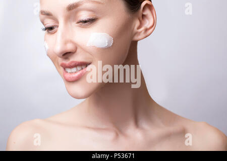 Lachende Mädchen Anwendung Feuchtigkeitscreme auf ihrem Gesicht. Foto der jungen Mädchen mit makelloser Haut auf grauem Hintergrund. Hautpflege und Schönheit Konzept Stockfoto