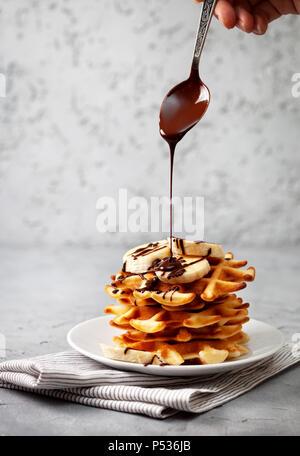 Belgische Waffeln mit Schokoladensauce und Banane auf grauem Beton Hintergrund Stockfoto