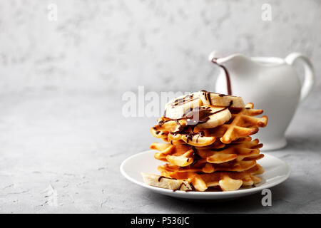 Belgische Waffeln mit Schokoladensauce und Banane auf grauem Beton Hintergrund Stockfoto