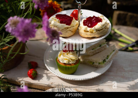 Scones mit Sahne und Marmelade auf Kuchen stand mit Sandwiches und Kuchen im Garten mit Blumen auf Kuchen Stand Stockfoto