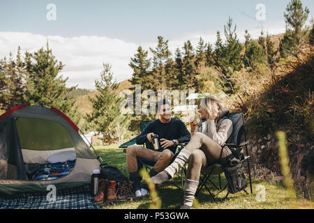 Der Mann und die Frau sitzen in Stühlen außerhalb des Zeltes Kaffee und Reden. Paar camping im Wald. Stockfoto