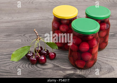 Drei Gläser mit eingemachten Kirschen. Prunus avium. Home Canning von reifen Beeren. Glasflaschen, fruchtig, Kompotte. Bündel von frischem Obst. Holz Hintergrund. Stockfoto