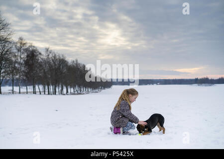 Kleine blonde kaukasischen Schwedische Mädchen spielen mit Hund im Winter Landschaft. Schönen kalten Abend bei Sonnenuntergang mit Schnee auf dem Feld und Lindenallee. Stockfoto