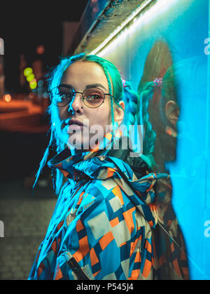 Junge hübsche Mädchen mit ungewöhnlichen Frisur in der Nähe von glühenden Türkis Neon Licht der Stadt bei Nacht. Blau gefärbte Haare zu Zöpfen. Nachdenklich hipster Jugendlicher in Gläser. Stockfoto
