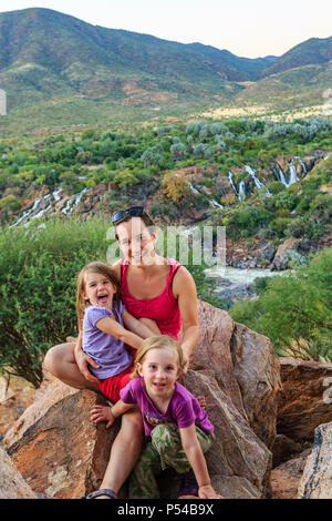Mutter mit zwei Mädchen sitzen auf den Felsen in der grünen Landschaft, hinter Epupa Wasserfälle, Kunene, Kunene Region, Namibia Stockfoto