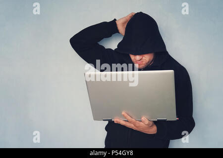 Nachdenklich Hacker mit seinem Computer denken auf blauen Hintergrund.. Hacker in einem schwarzen Haube mit einem grauen modernen ultrabook. Identitätsdiebstahl. Stockfoto