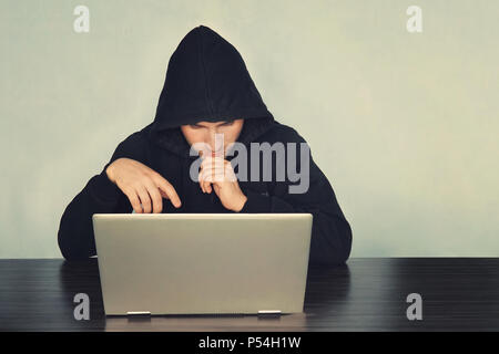 Schwarz vermummte Hacker im schwarzen T-Shirt auf dem Schreibtisch vor dem Laptop sitzt und Denken somethings. Er ist auf der Suche nach Laptop. Blur ba Stockfoto